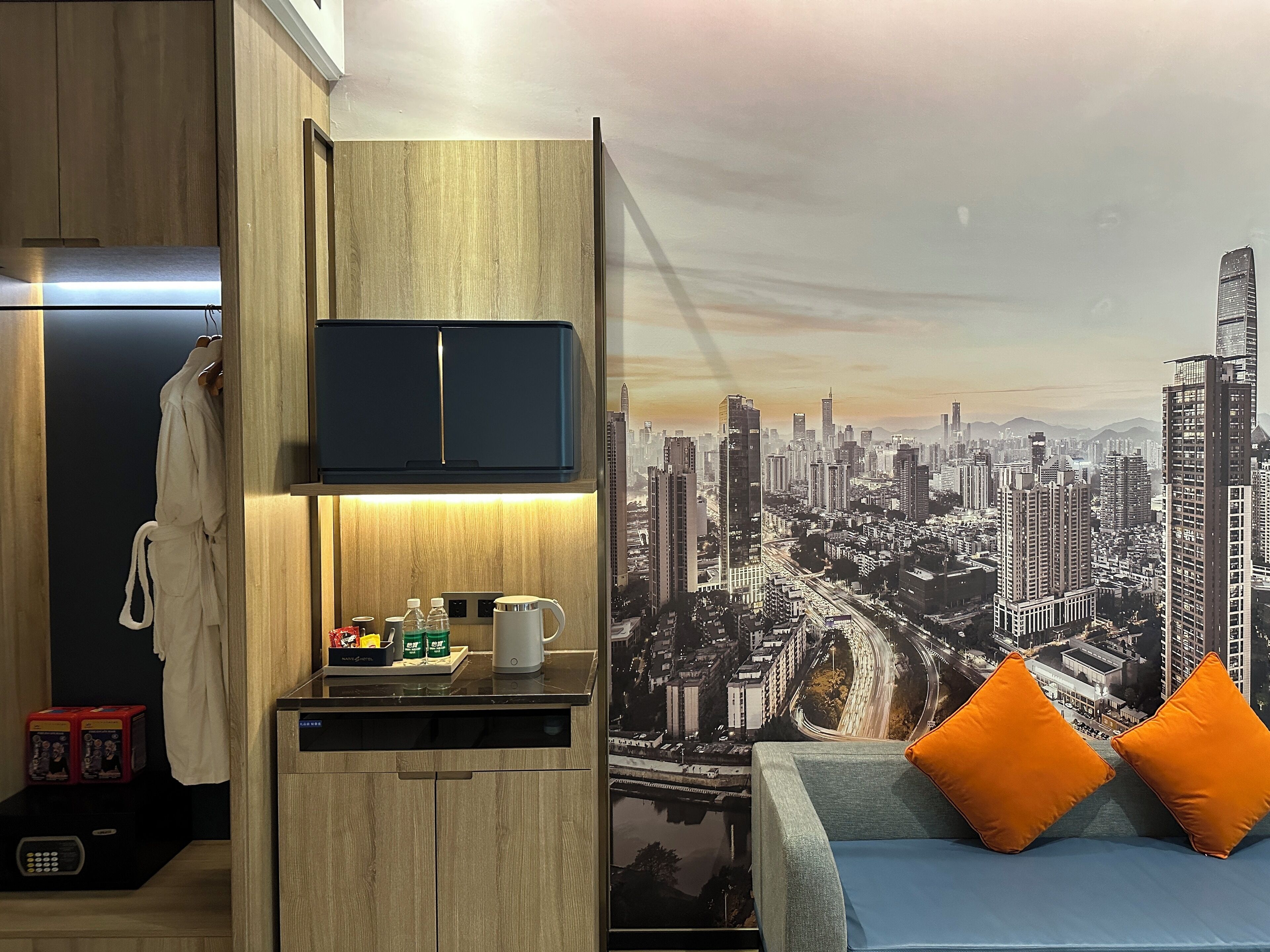 Habitación compacta con un mural de la ciudad, microondas integrado, una pequeña mesa de centro y una cama con almohadas naranjas.