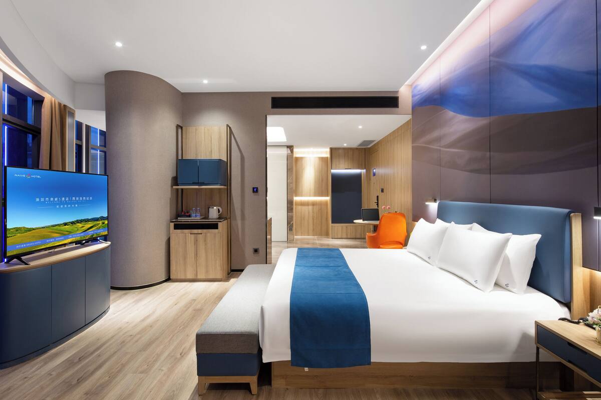 A modern hotel room with a large bed, a TV, a desk, and a bathroom visible in the background.