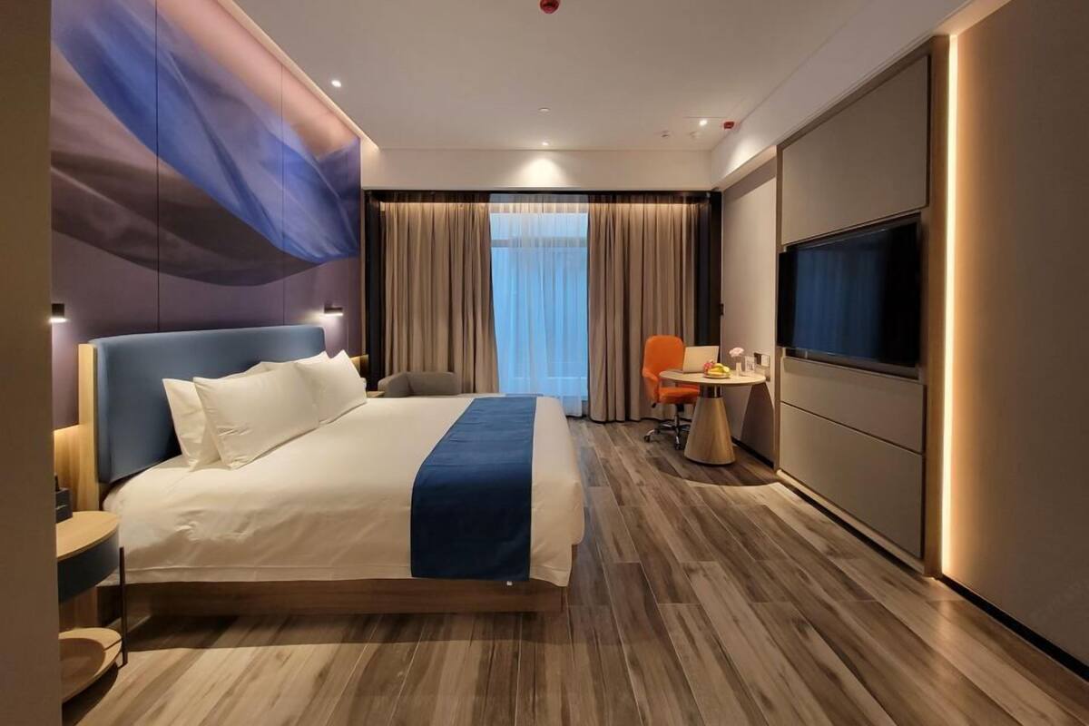 A modern hotel room with a large bed, a desk with a chair, a TV, and a large window with curtains.