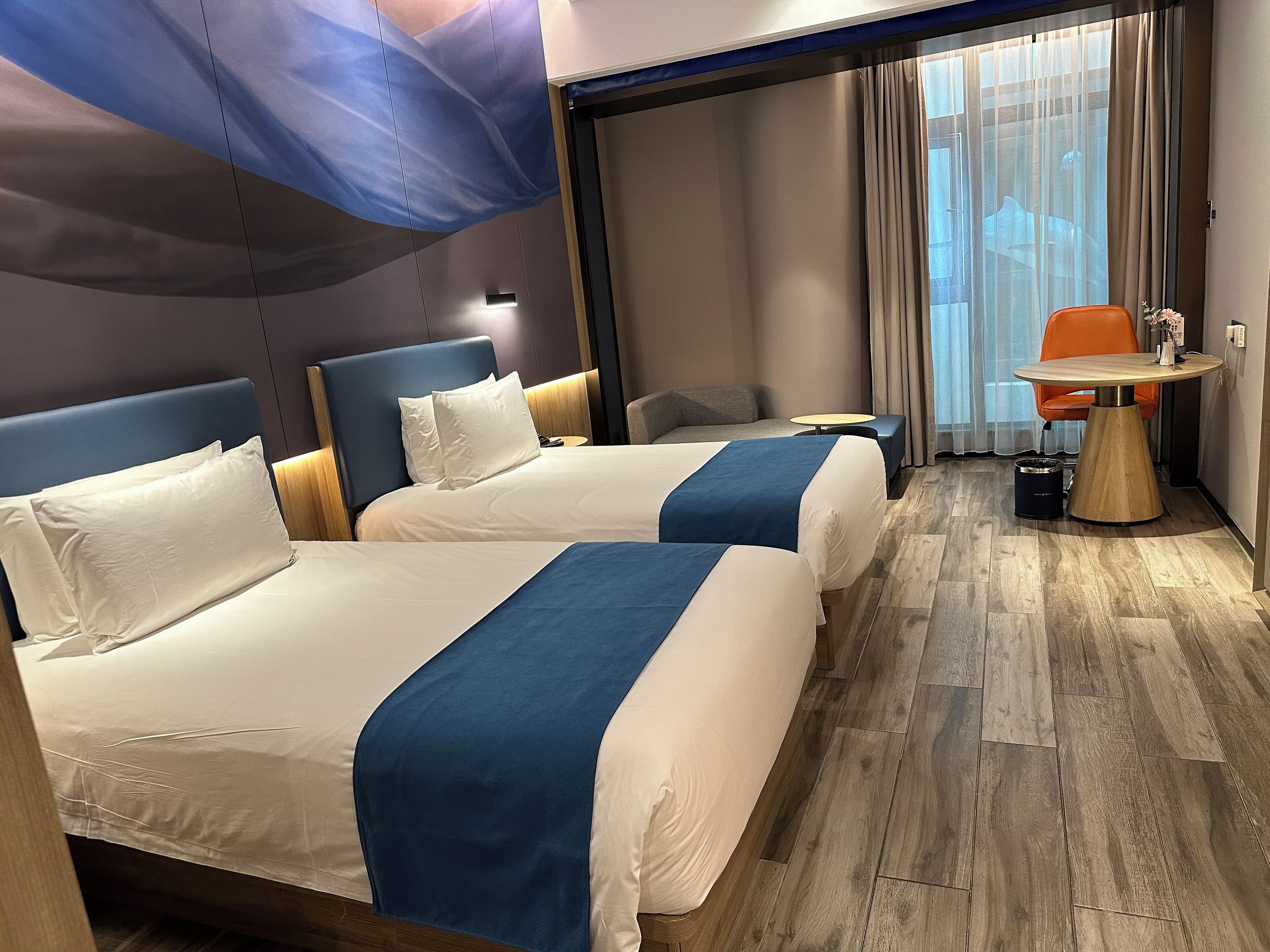 A hotel room with two beds, a small table, a chair, and a view of the outdoors.