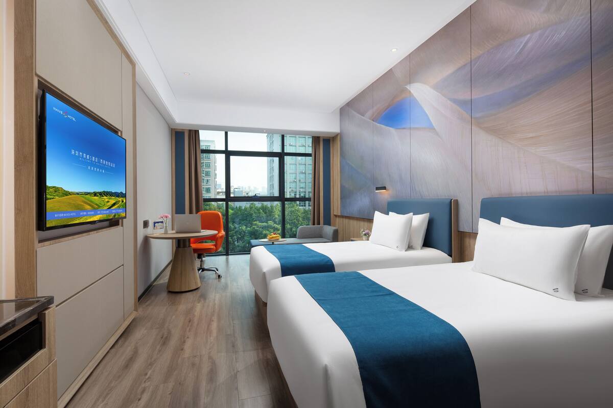 A hotel room with two beds, a desk with a chair, a TV on the wall, and a large window with a city view.