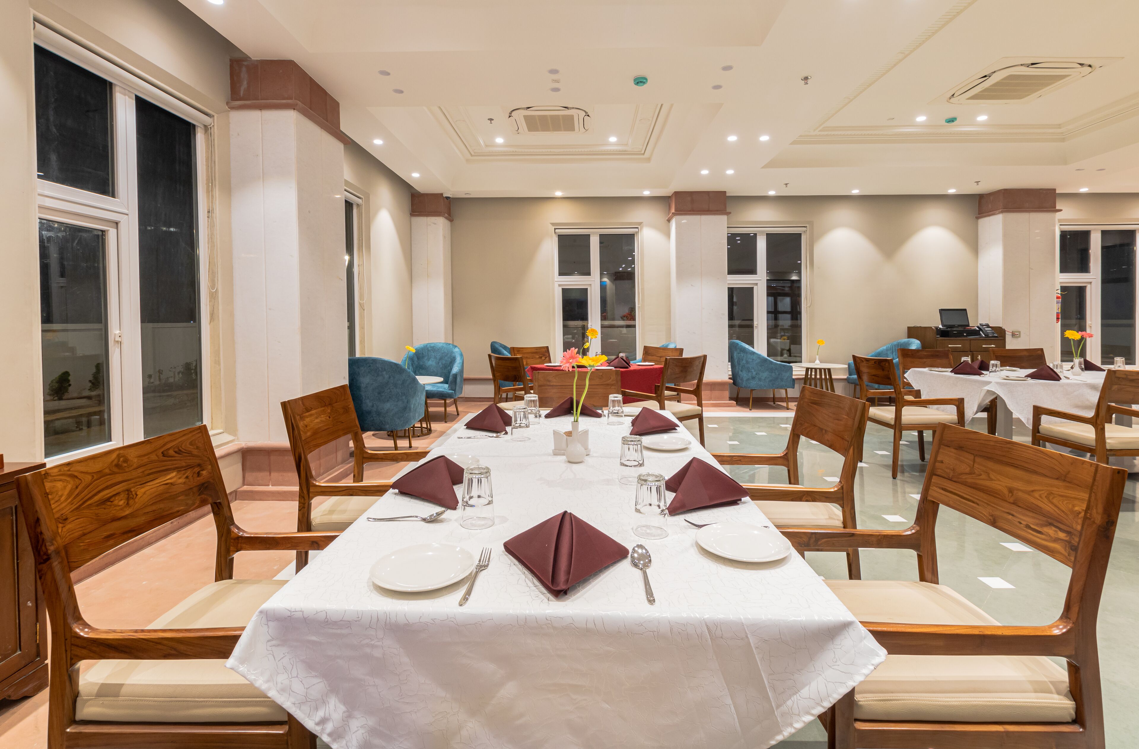 Un comedor con varias mesas dispuestas para una comida, sillas de madera y vistas al exterior a través de amplios ventanales.