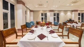 Un comedor con varias mesas dispuestas para una comida, sillas de madera y vistas al exterior a través de amplios ventanales.