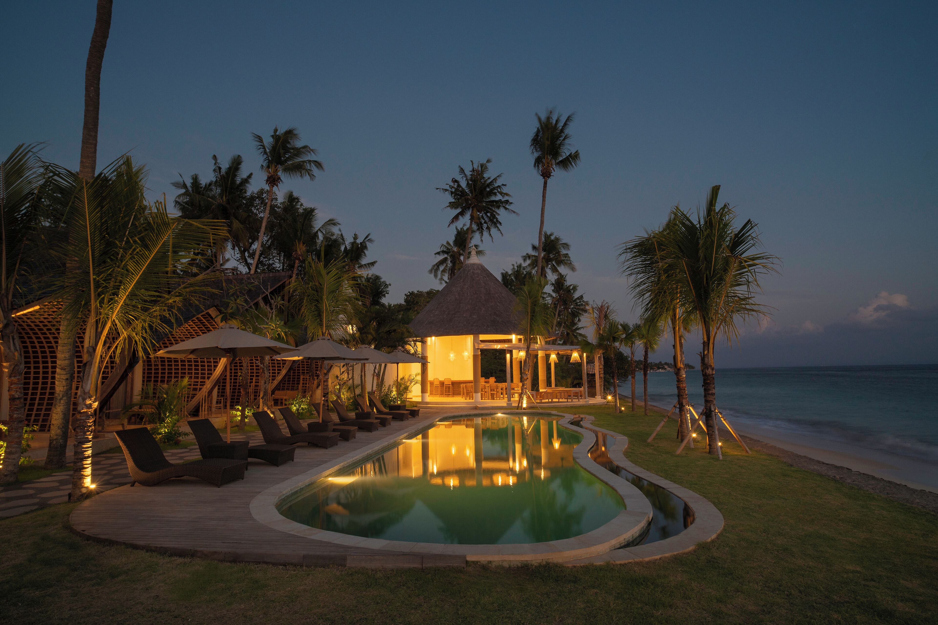 A poolside area with lounge chairs and umbrellas, a thatched-roof structure, and palm trees.