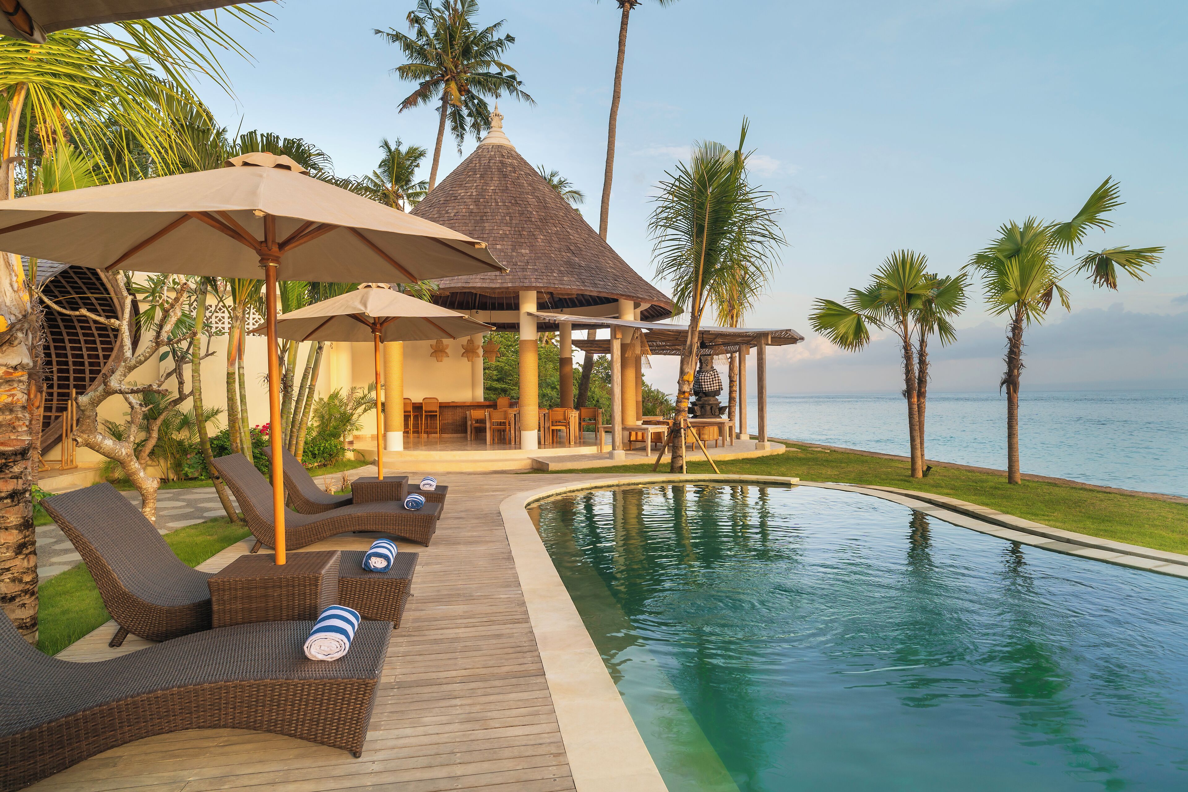 Un espace aménagé au bord de la piscine, avec des chaises longues et des parasols, un coin salon abrité et des palmiers.