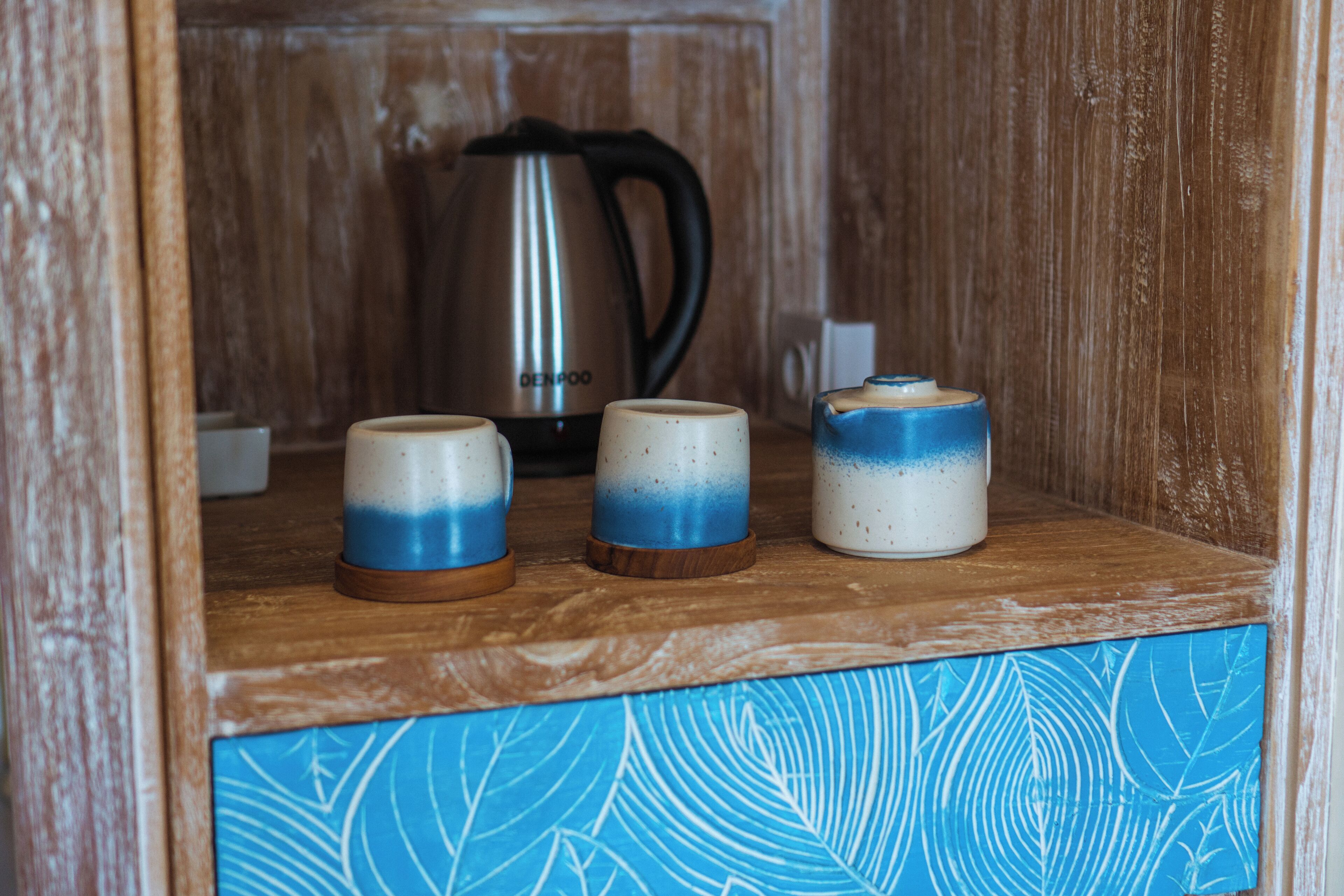 A wooden shelf with three ceramic mugs and a stainless steel kettle.