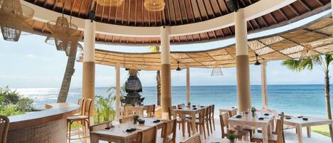 A restaurant with a view of the ocean, featuring wooden tables and chairs, a bar area, and a thatched roof.