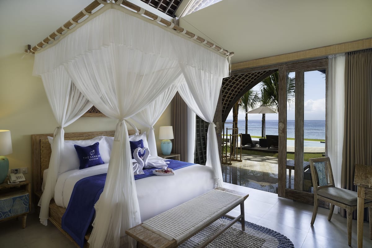 A four-poster bed with a canopy, a bedside table with a lamp, and a view of the ocean through a glass door.