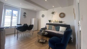 A living room with a sofa, chairs, a coffee table, a refrigerator, and a window with a view of buildings. - La vie est belle - Arromanches center, terrace (Arromanches-les-Bains)