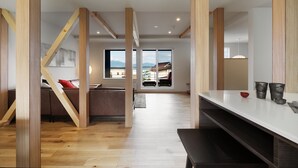 A modern interior with exposed wooden beams, a dining area with a white table, and a view of a residential building through large windows. - AKATSUKI (Furano)