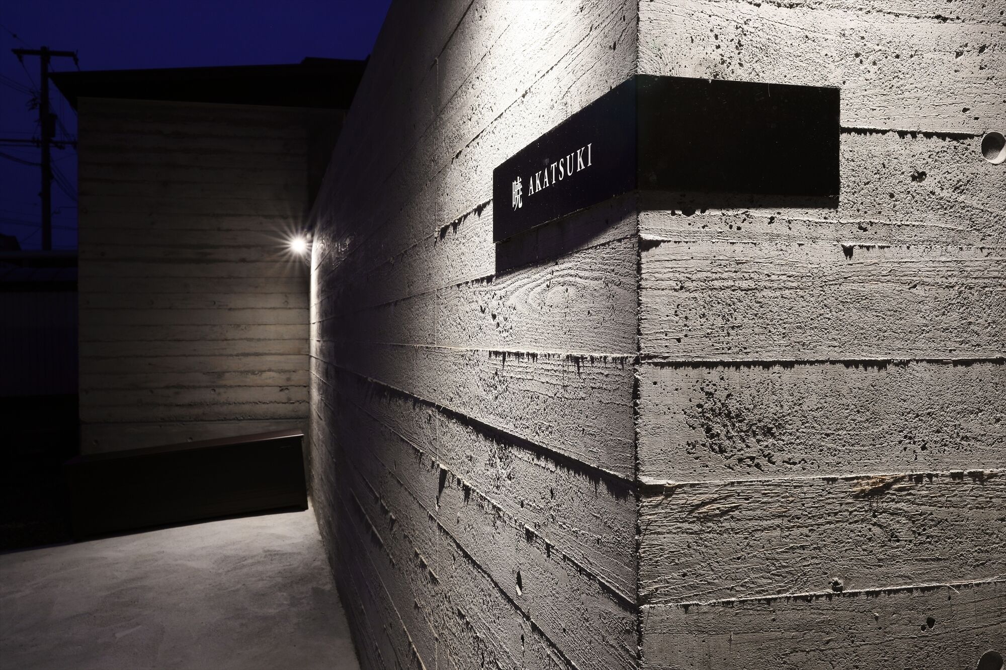 Un muro de ladrillo con un cartel que dice "AKATSUKI" y una fuente de luz a la izquierda.