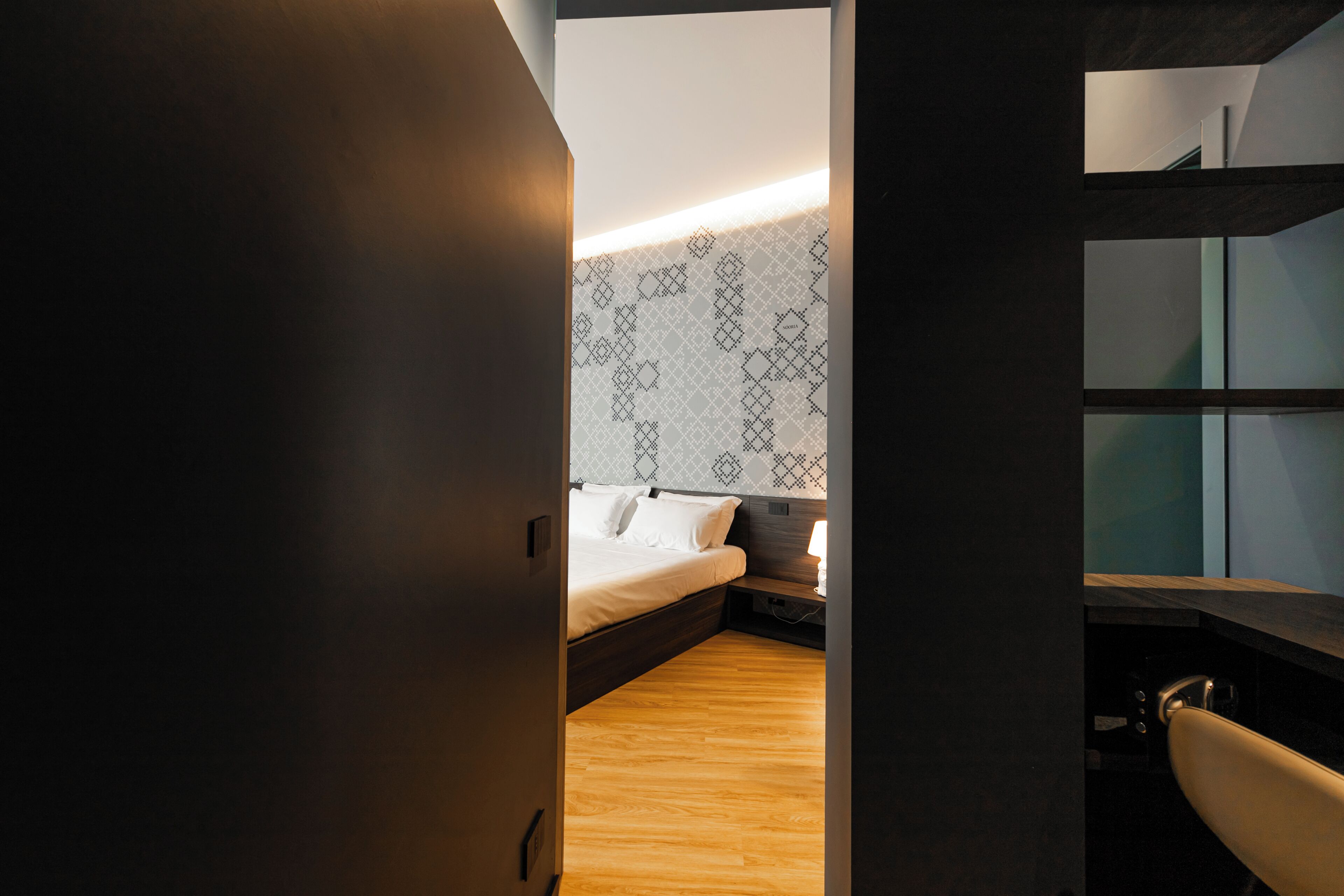 A modern bedroom with a wooden floor, a bed with white linens, a patterned wall, and a desk with a chair.