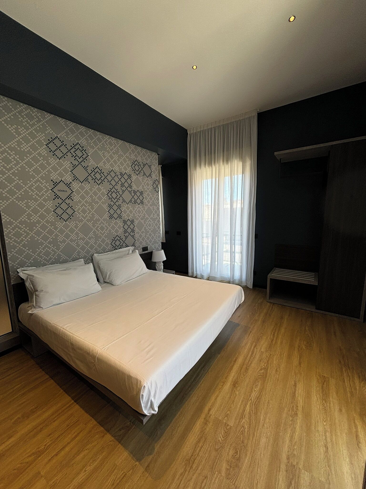 Una cama grande en una habitación moderna con cabecera de diseño geométrico, una mesita de noche con lámpara y una ventana con cortinas translúcidas.