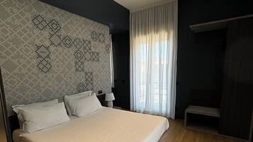 Una cama grande en una habitación moderna con cabecera de diseño geométrico, una mesita de noche con lámpara y una ventana con cortinas translúcidas.