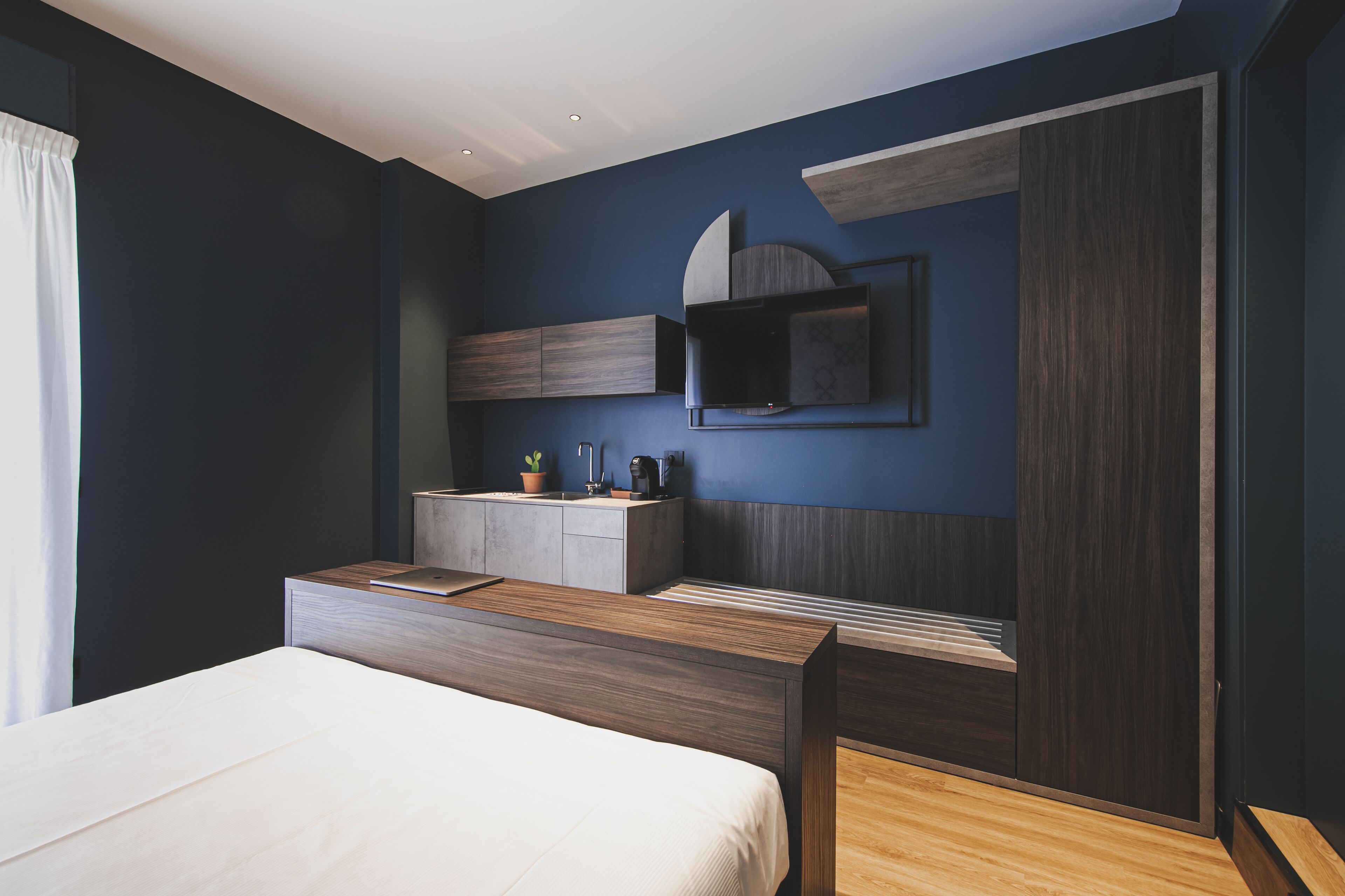 A modern bedroom with a bed, bedside table, built-in wardrobe, and a small kitchenette area.