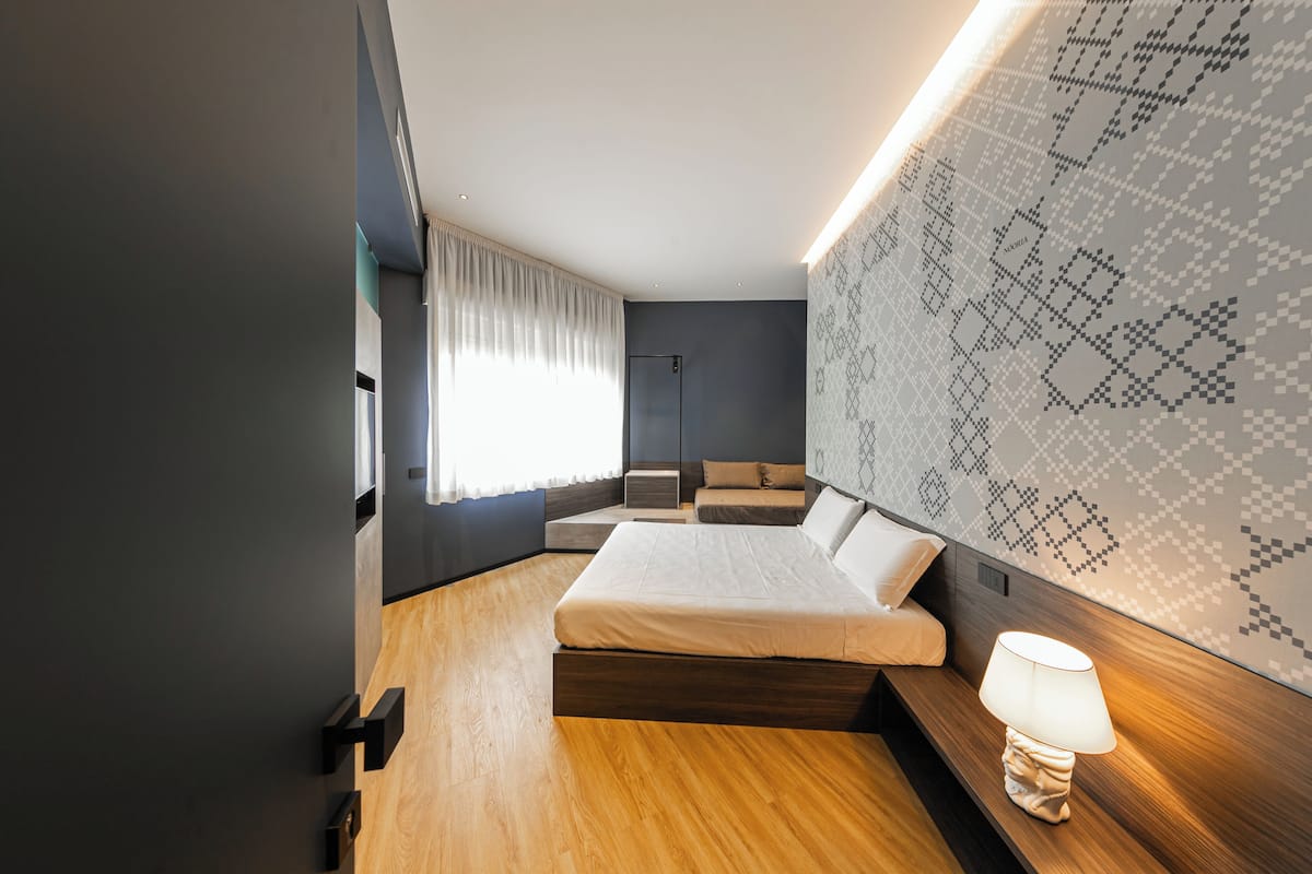 Ein modernes Schlafzimmer mit Holzboden, einem Bett mit weißer Matratze, einem Nachttisch mit Lampe, einem großen Fenster mit Vorhängen und einer Wand mit Muster.
