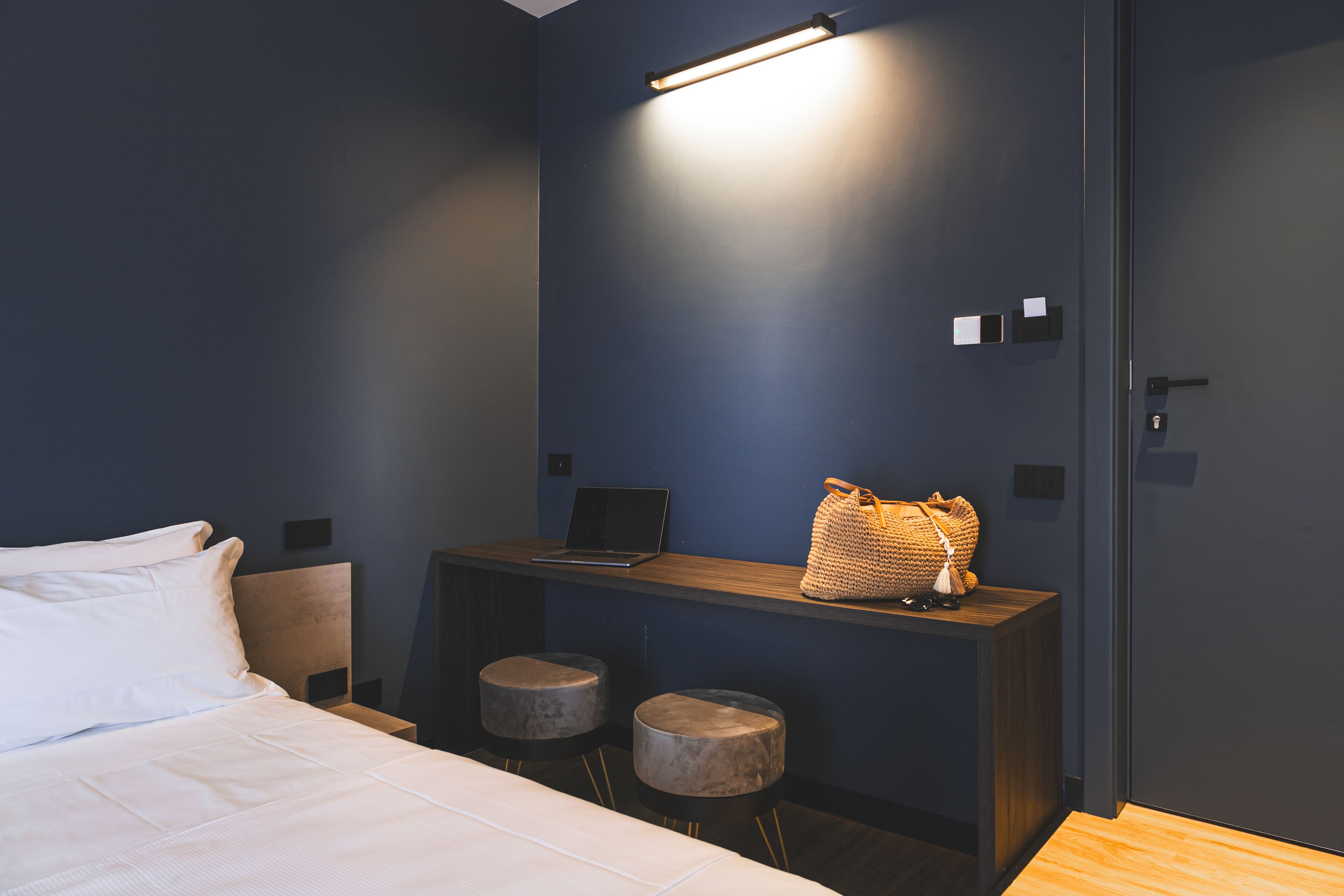 A modern hotel room with a bed, a desk with a laptop, two stools, and a wall-mounted light fixture.
