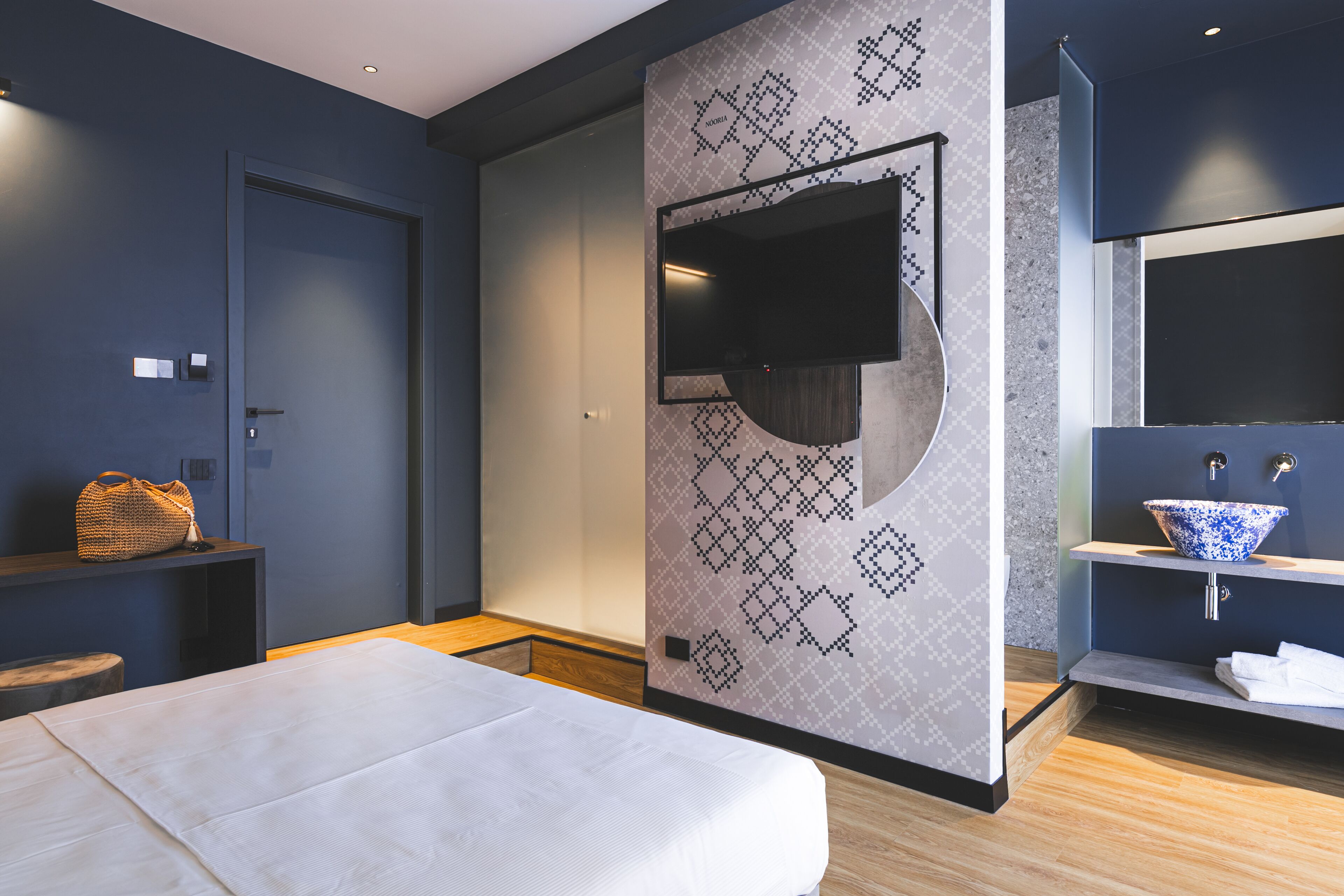 A modern hotel room with a bed, a TV mounted on a geometric-patterned wall, a wooden floor, and a bathroom with a blue and white sink.