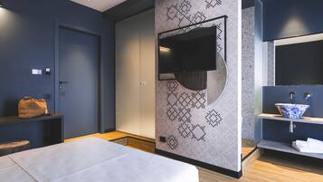 A modern hotel room with a bed, a TV mounted on a geometric-patterned wall, a wooden floor, and a bathroom with a blue and white sink.