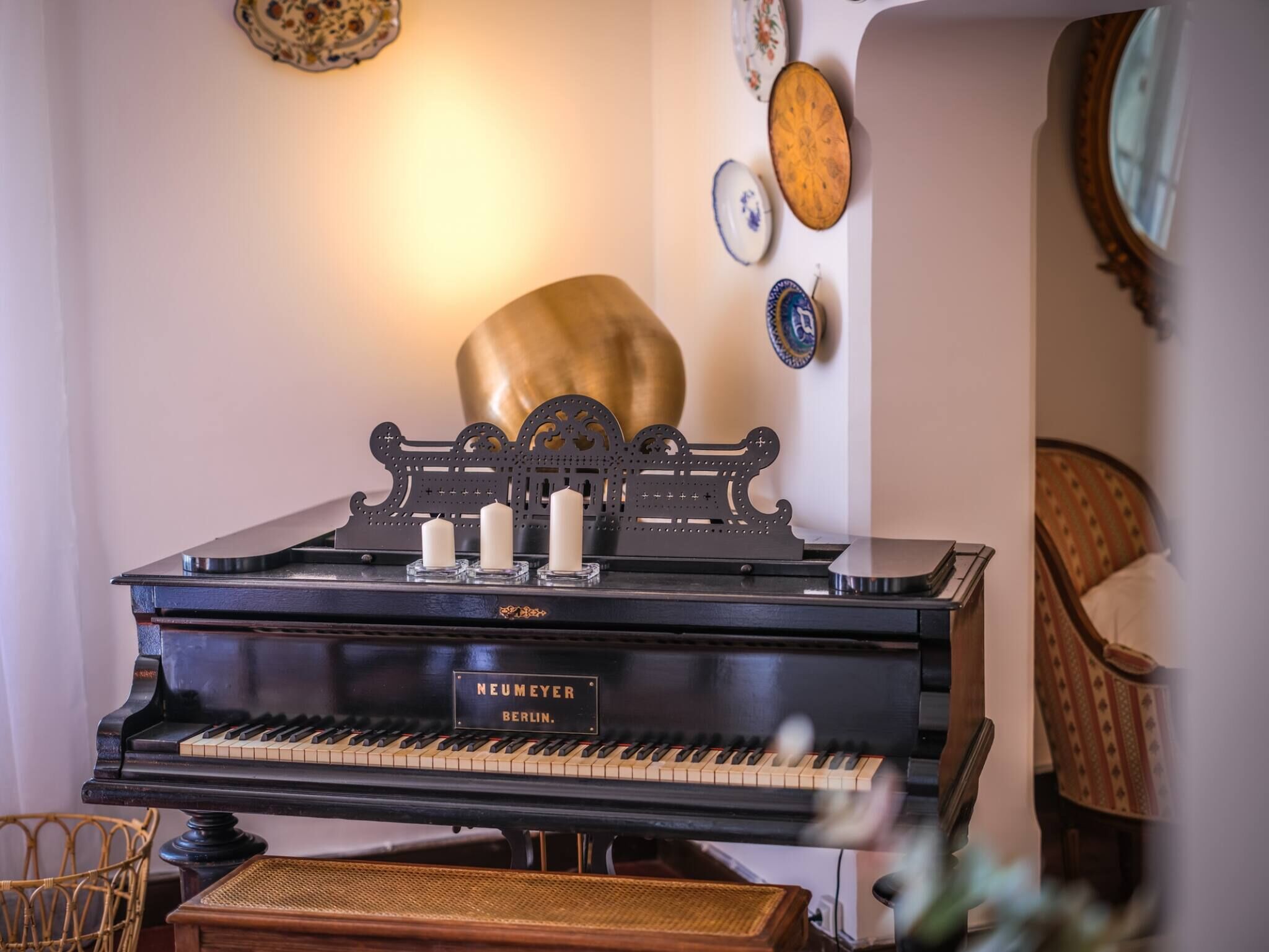 Un piano à queue sur lequel sont posées trois bougies blanches, un mur décoratif orné de différentes assiettes, et une chaise en osier au premier plan.