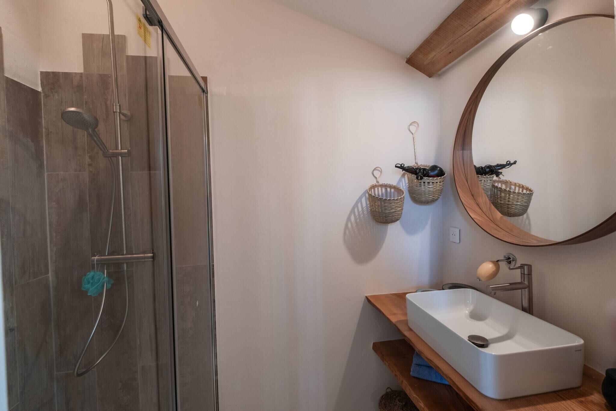 A bathroom with a glass-enclosed shower, a wooden vanity with a white sink, and a mirror with baskets on the wall.