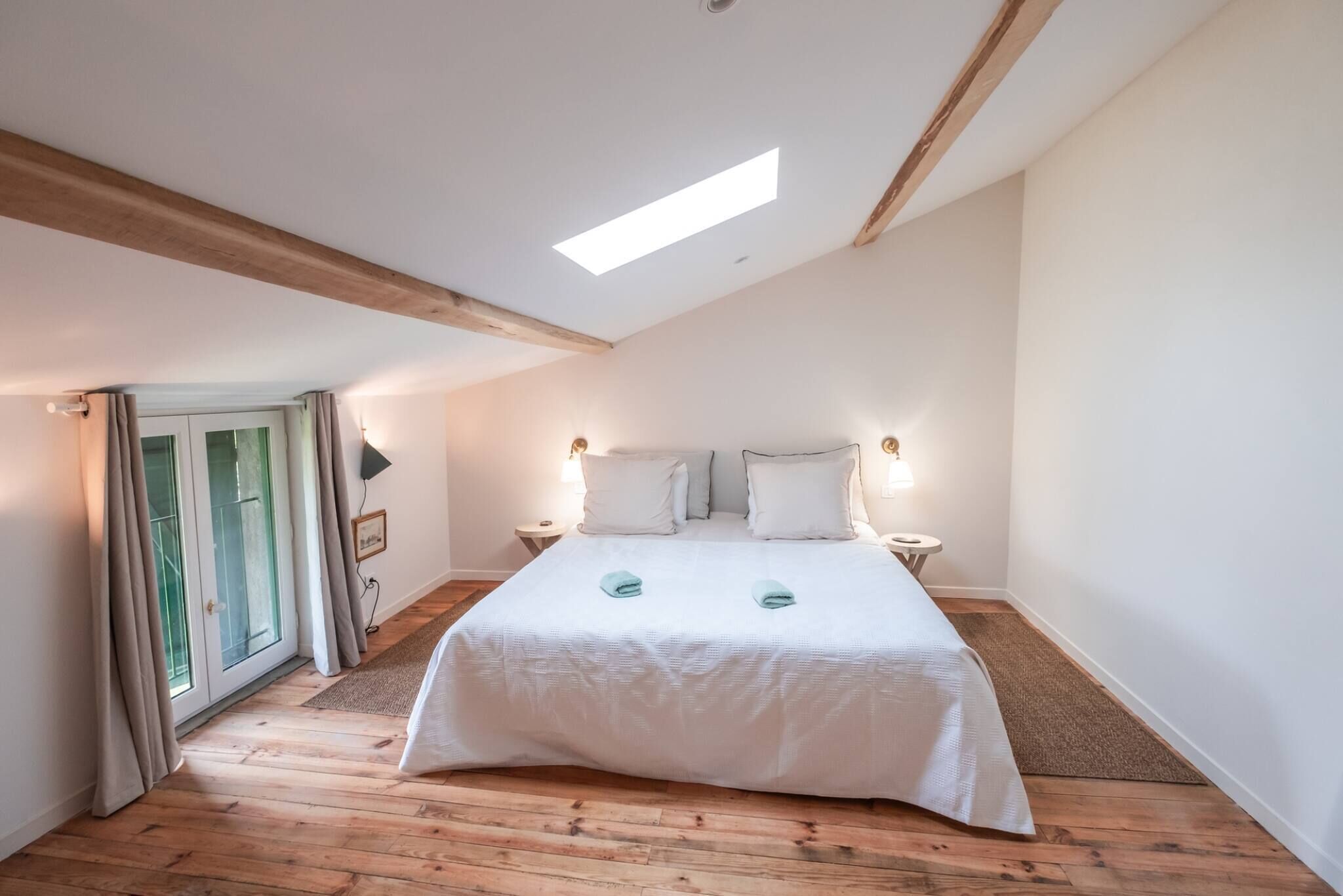 A bedroom with a large bed, wooden beams, a skylight, and a view through a glass door.