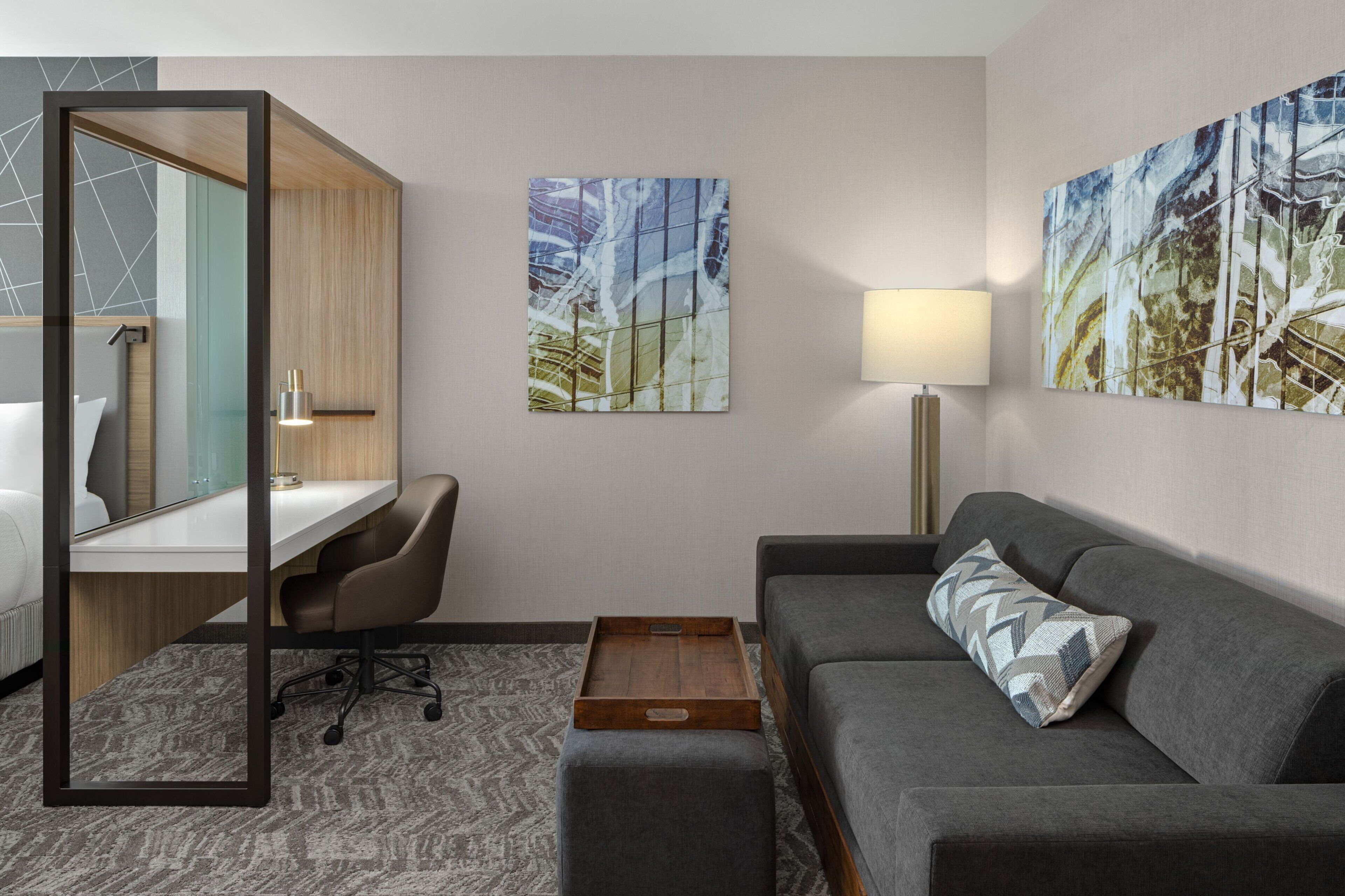 A hotel room with a desk, a grey sofa, a small wooden table, and two abstract paintings on the walls.