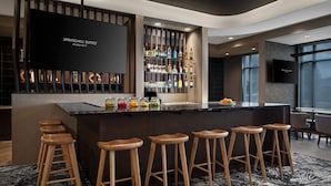 A bar area with a black countertop, wooden stools, and a variety of bottles and glasses. - Springhill Suites By Marriott Kalamazoo Portage (Portage)