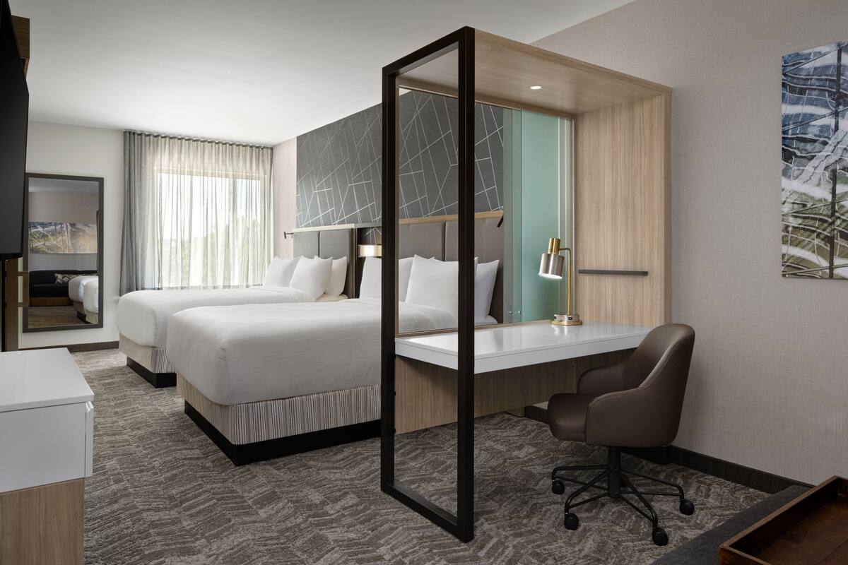 A hotel room with two beds, a desk, a chair, a mirror, and a painting on the wall.