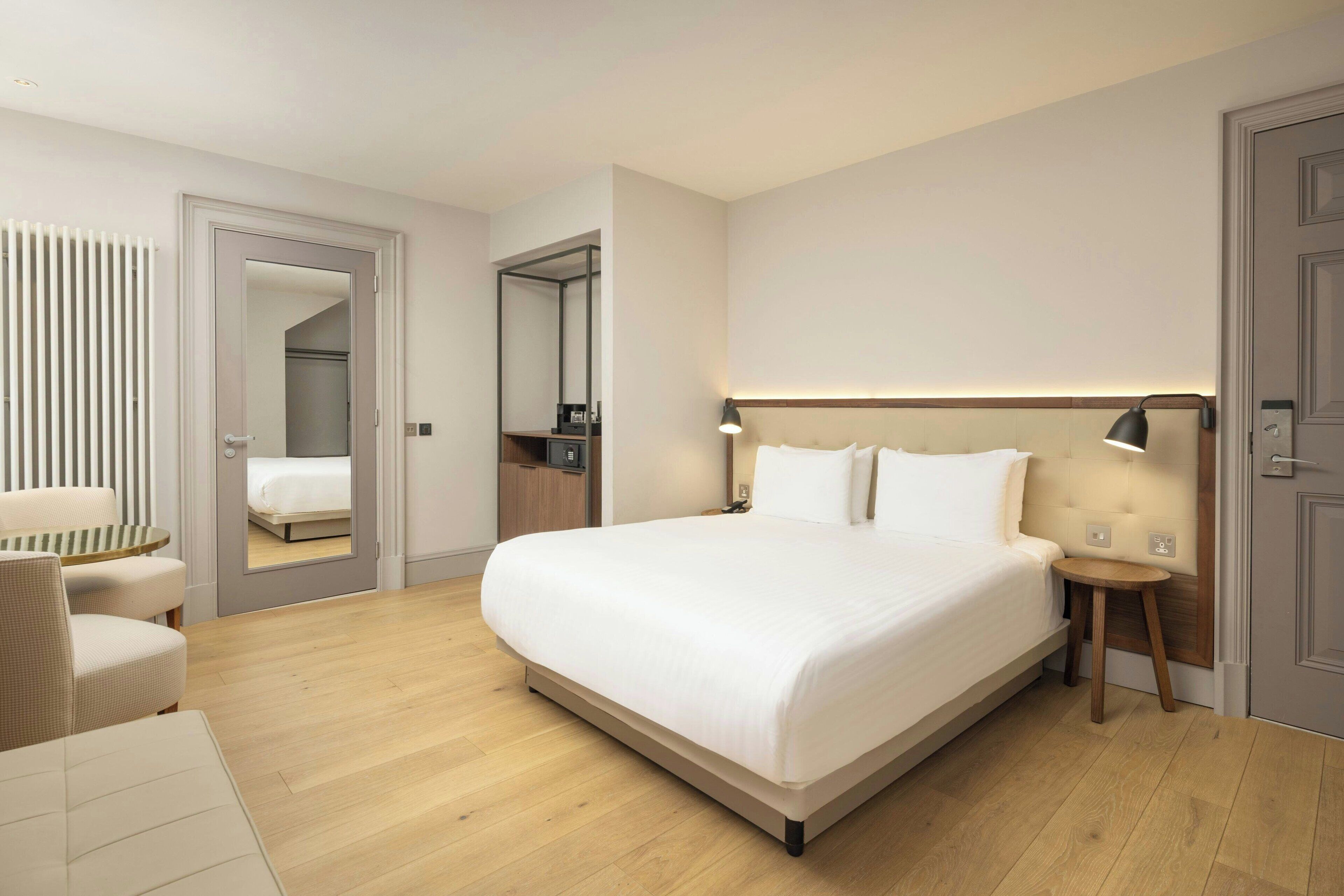 Una habitación de hotel moderna con una cama grande, una mesita de noche, una silla, un escritorio, un espejo y un armario.