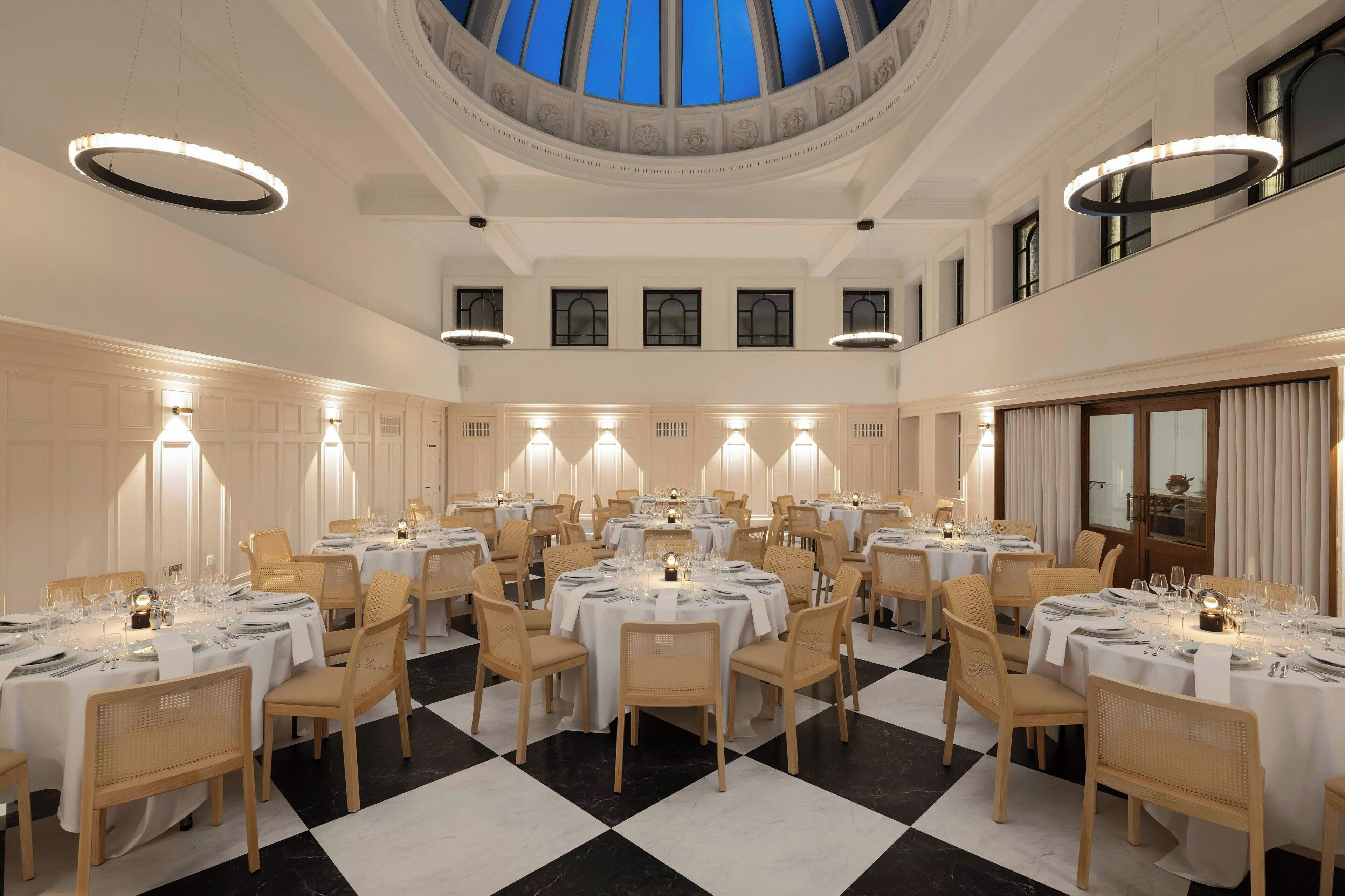 A large dining hall with multiple round tables, white tablecloths, and wicker chairs.