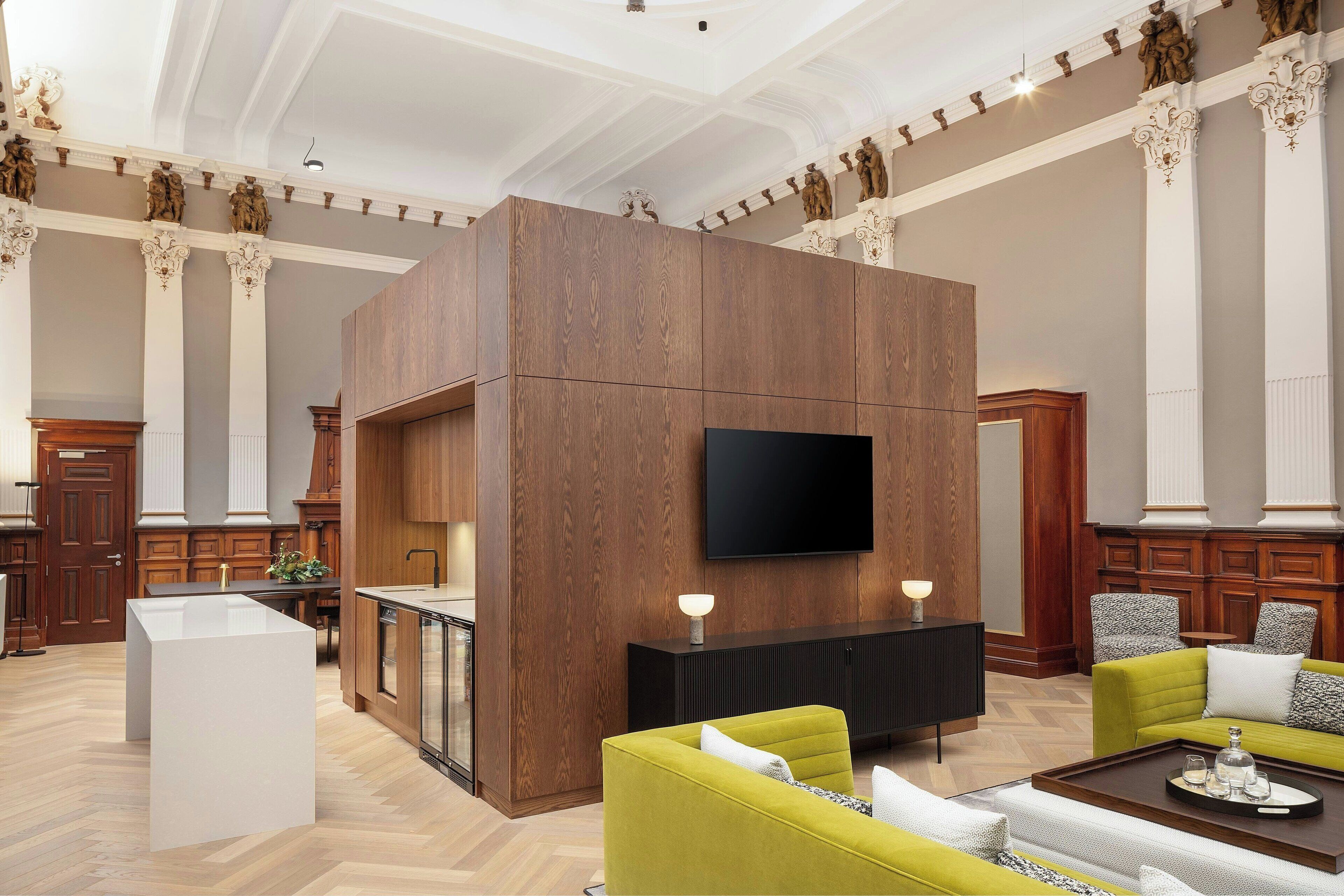 Un hall d’entrée spacieux avec un coin cuisine moderne, un téléviseur à écran plat fixé à un mur en bois et un espace salon avec des canapés verts.