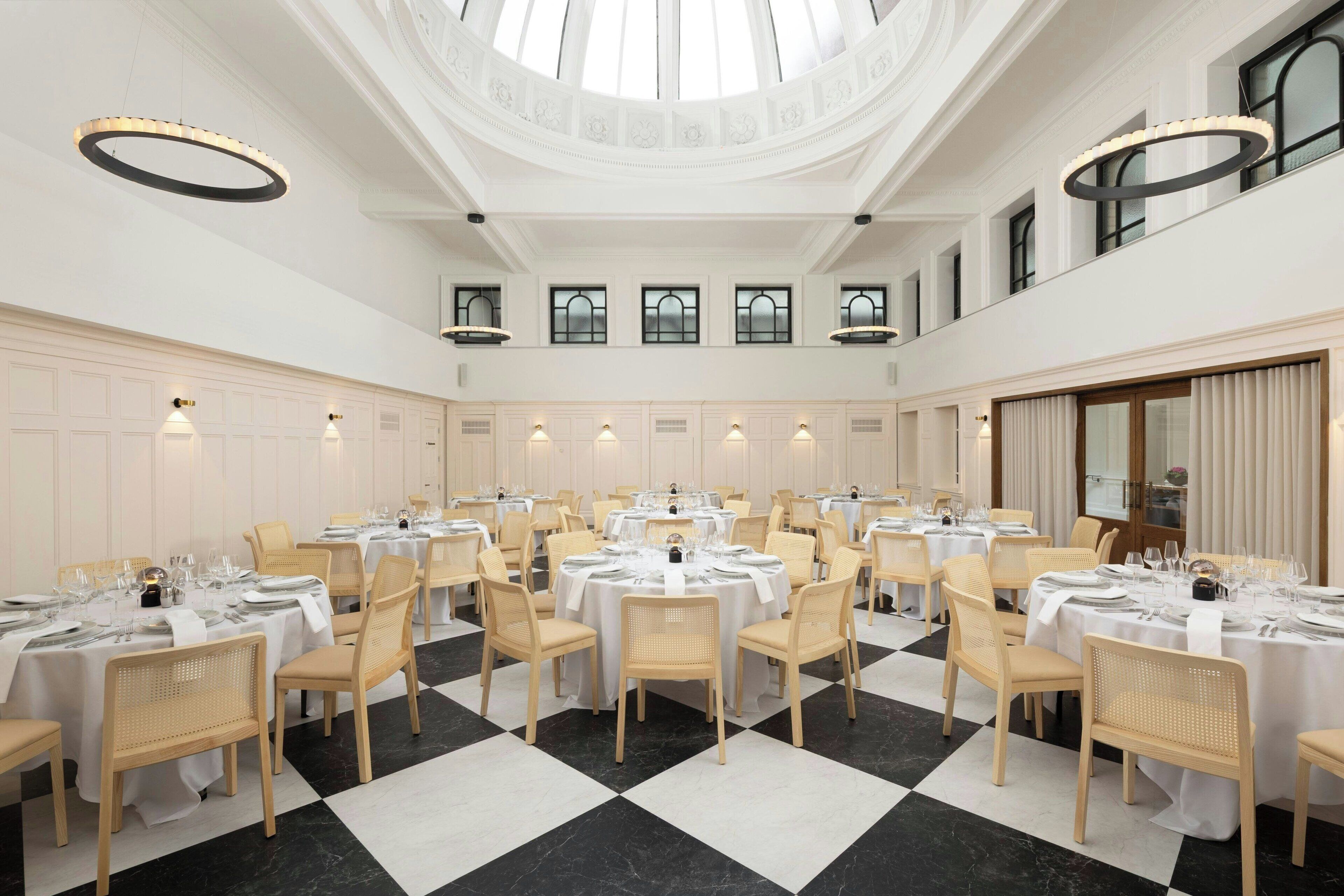 A spacious dining hall with multiple round tables, white tablecloths, and light-colored chairs.