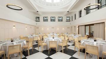 A spacious dining hall with multiple round tables, white tablecloths, and light-colored chairs.