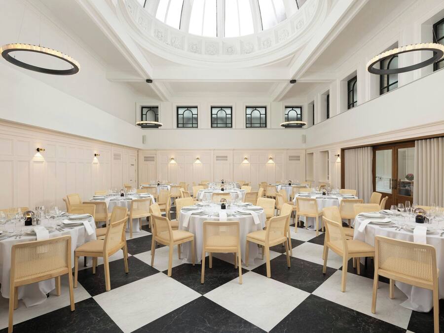 A spacious dining hall with multiple round tables, white tablecloths, and light-colored chairs.