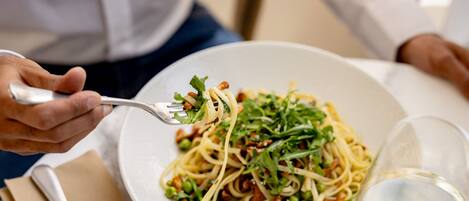 Un plato de pasta con guisantes y verduras, un tenedor, una copa de vino blanco y una servilleta sobre una mesa.