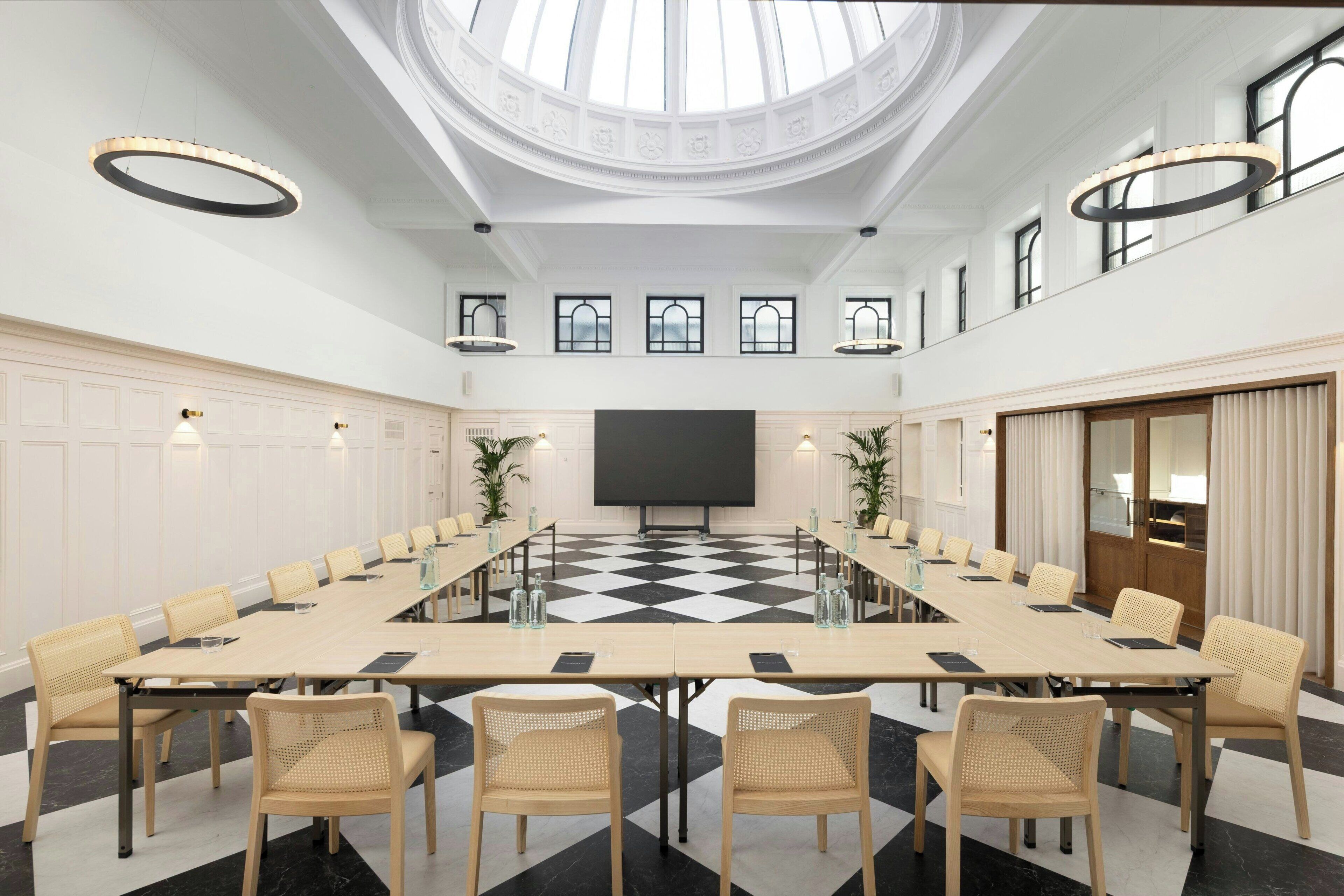 Een vergaderzaal met een lange tafel, stoelen, een groot scherm en een geruite vloer.