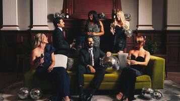 A group of people seated on a green sofa with disco balls and champagne bottles in a room with classical architecture.