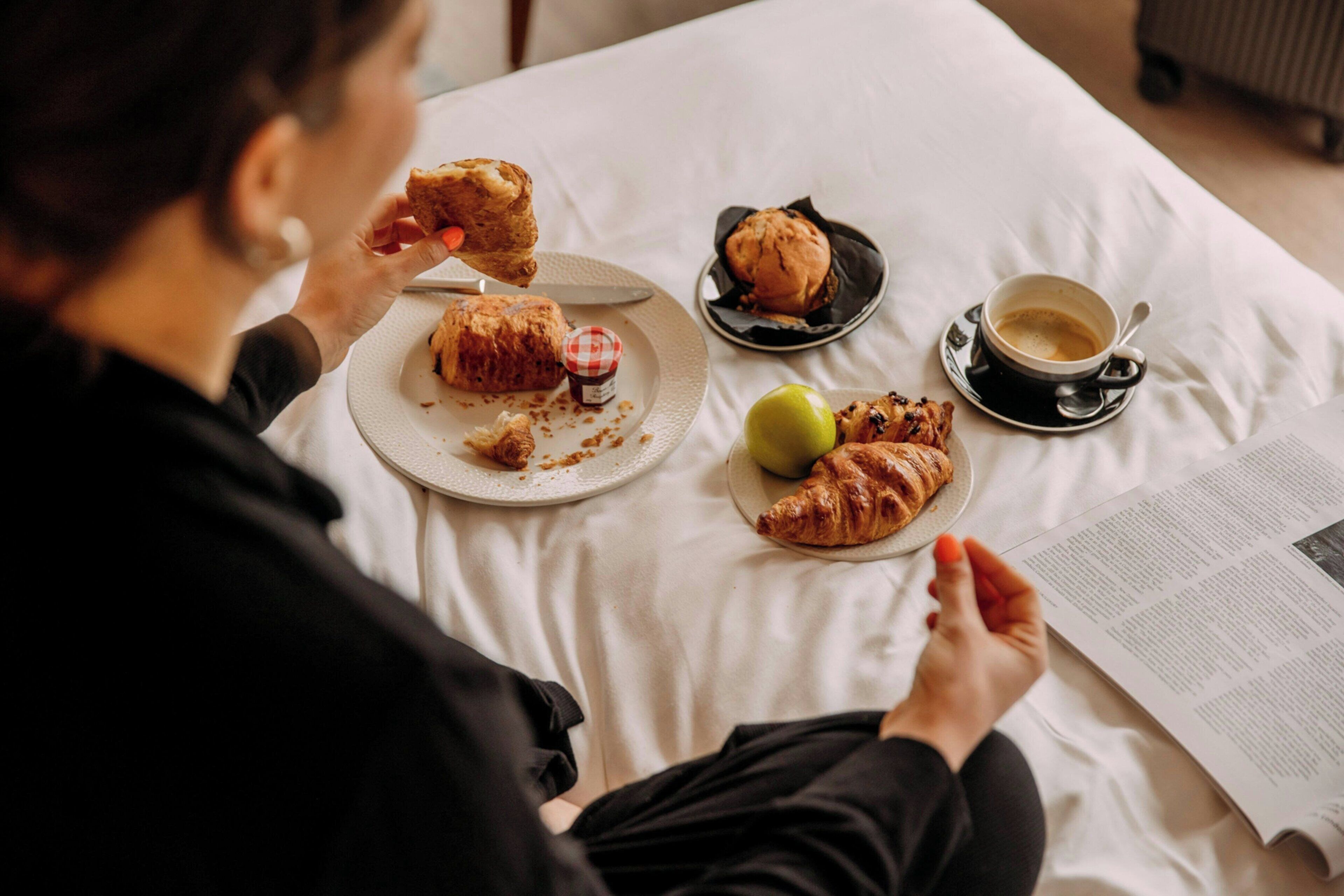 A person sitting on a bed with a plate of food, a croissant, an apple, and a cup of coffee.