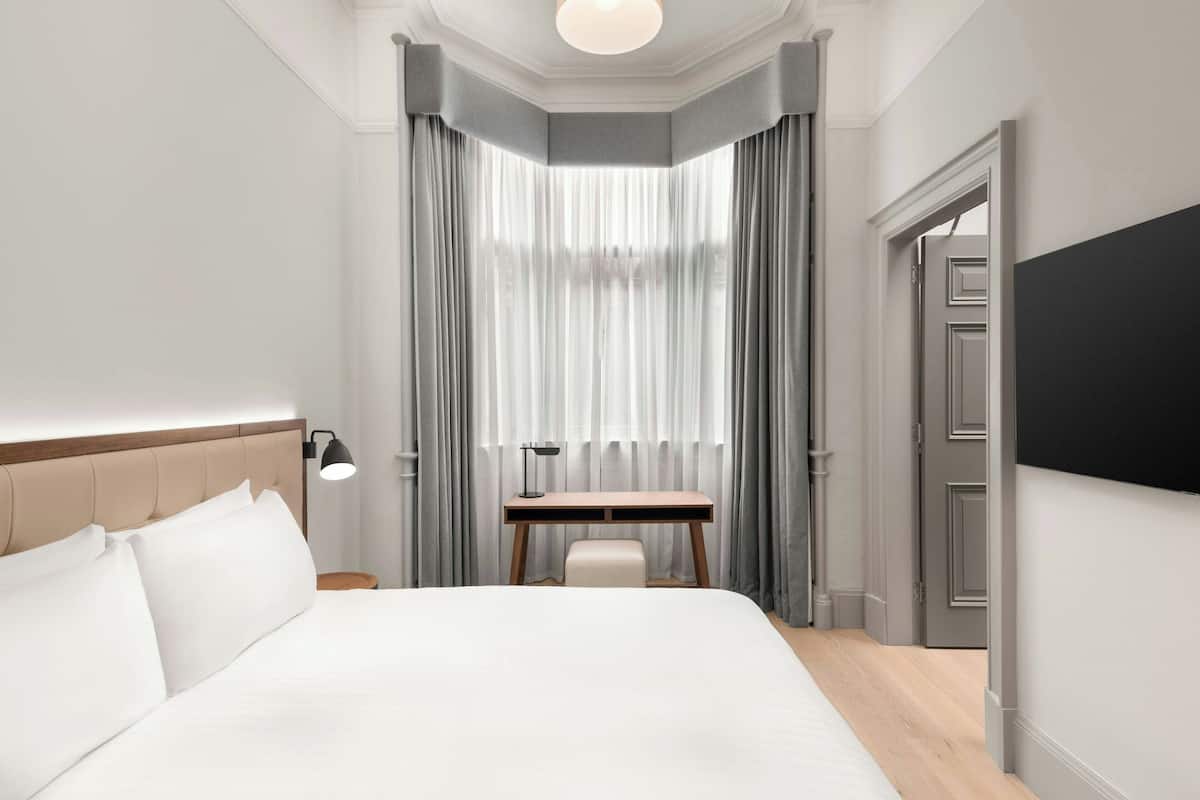 Ein Hotelzimmer mit einem großen Bett, einem Schreibtisch und einem Sessel. Es gibt ein Fenster mit Vorhängen, eine Tür und einen an der Wand montierten Fernseher.
