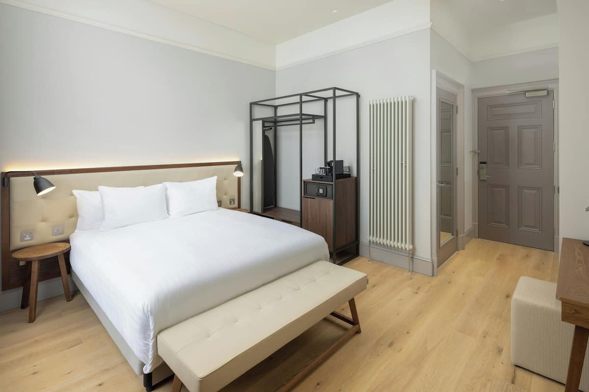 Ein modernes Hotelzimmer mit einem großen Bett, Nachttischen, einer Bank, einem Kleiderschrank und einem Heizkörper.