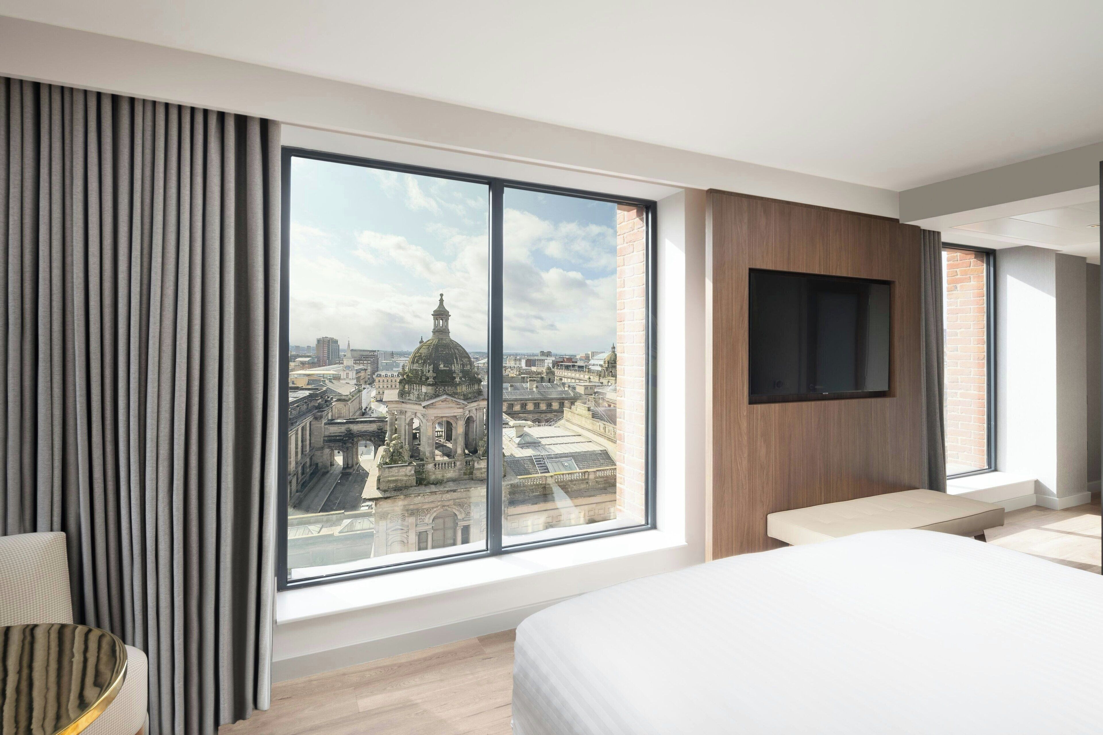 A modern hotel room with a large window offering a city view, a flat-screen TV mounted on the wall, and a bed with a wooden headboard.
