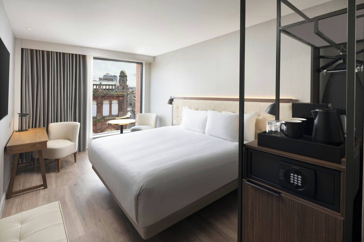 Ein Hotelzimmer mit einem großen Bett, einem Schreibtisch, einem Sessel, einer Kaffeemaschine und Blick auf ein historisches Gebäude.