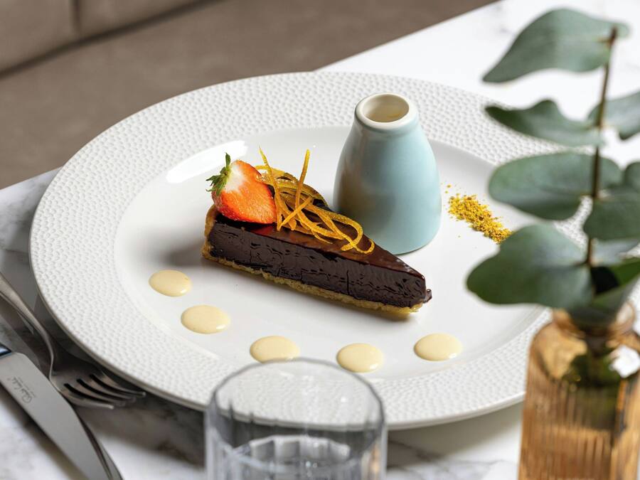 A dessert plate with a chocolate tart, a strawberry, and a small blue cup, accompanied by a glass of water and a vase with a plant.