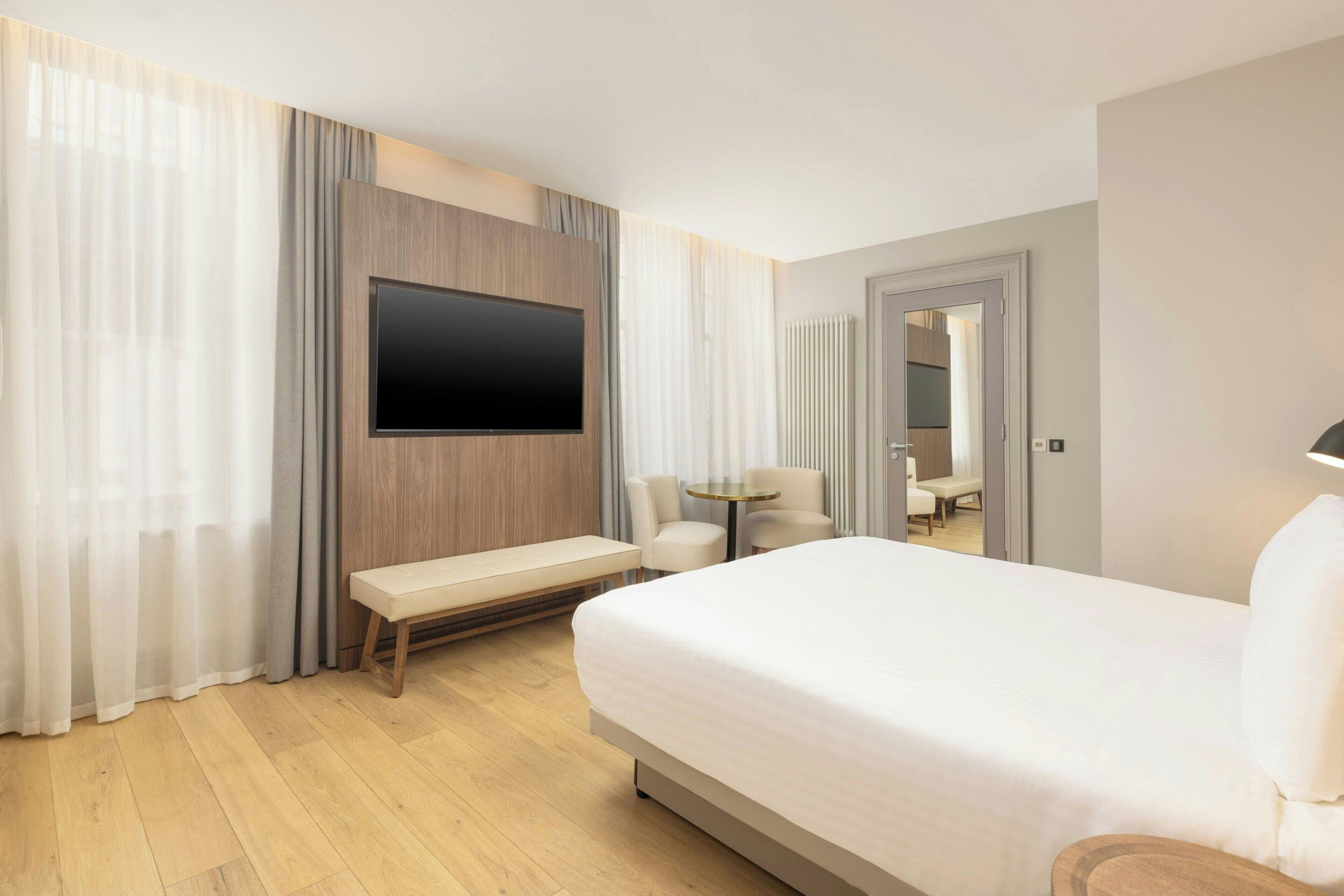 A hotel room with a large bed, a flat-screen TV mounted on the wall, a small round table, and a bench.