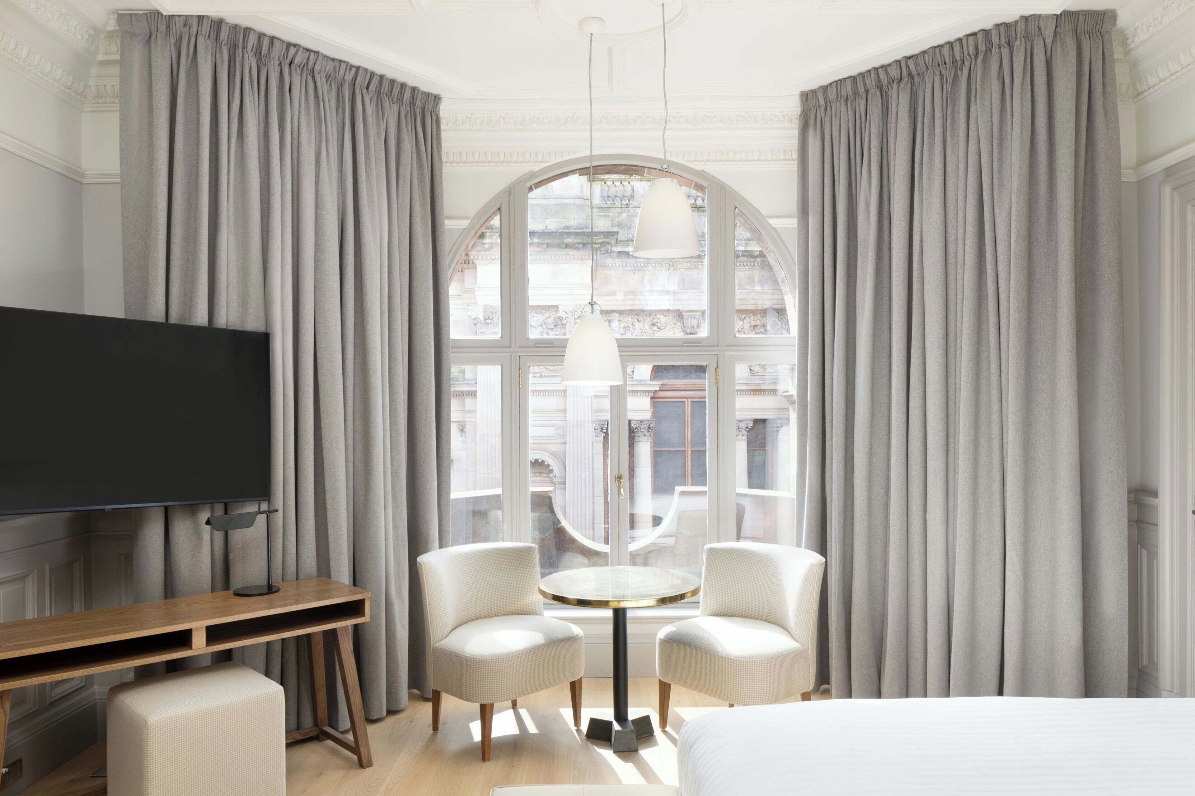 A hotel room with a large window, a flat-screen TV, a round table with two chairs, and a bed.