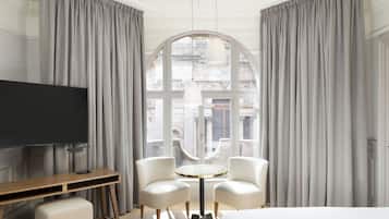 A hotel room with a large window, a flat-screen TV, a round table with two chairs, and a bed.