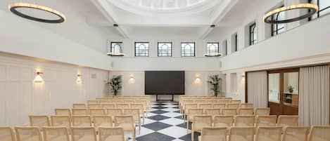 A conference room with a checkered floor, rows of chairs, a large screen, and a high ceiling with a skylight.