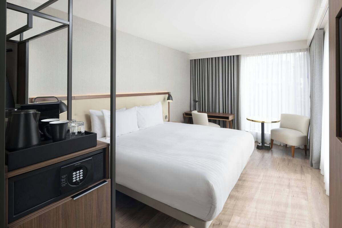 Ein Hotelzimmer mit einem großen Bett, einem Schreibtisch mit Stuhl, einer Kaffeemaschine und einem kleinen Tisch mit Lampe.