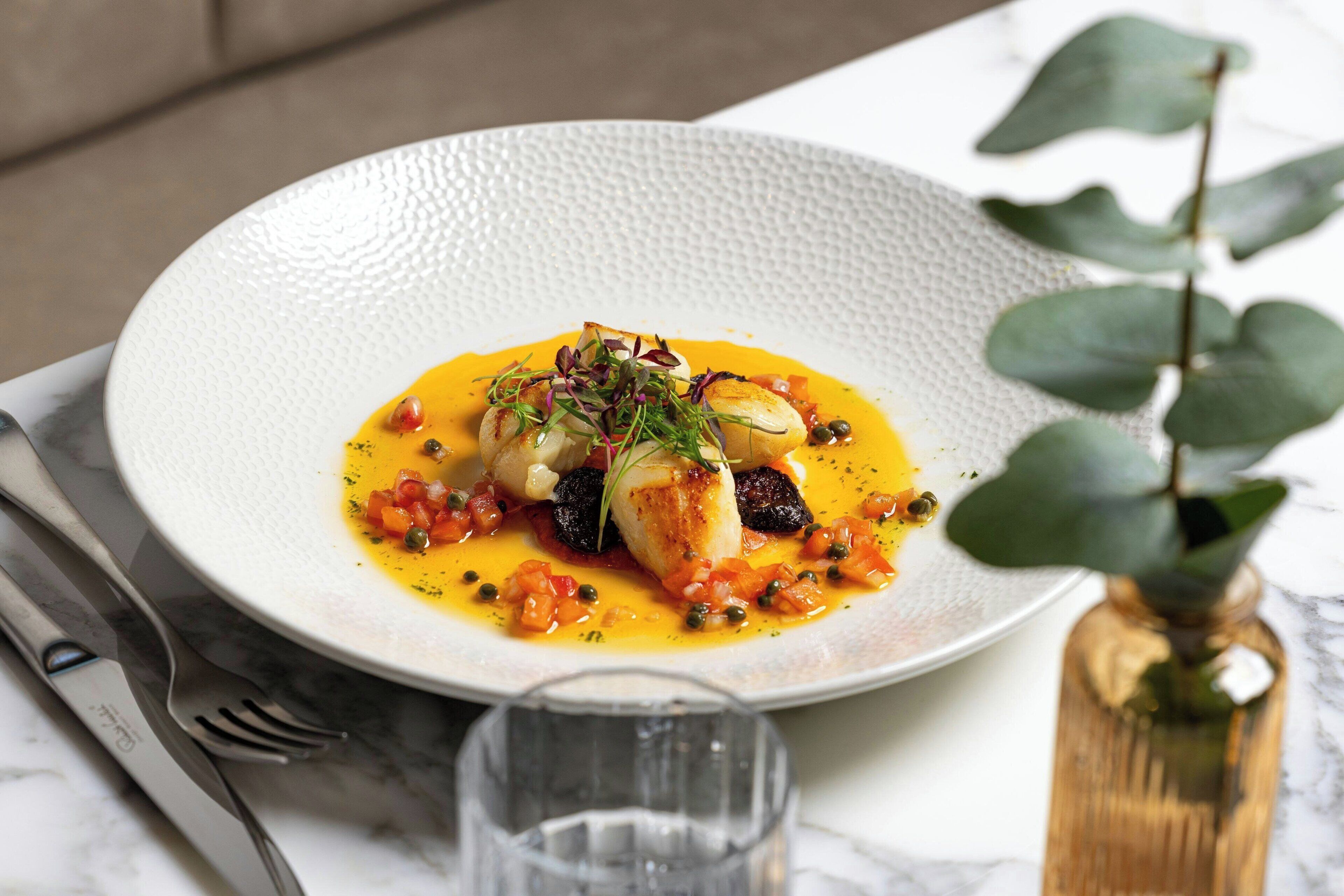 A white textured plate with a seafood dish, a glass of water, and a small vase with a plant on a marble table.